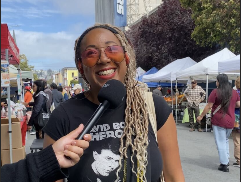 A smiling woman wearing sunglasses and a "David Bowie Heroes" t-shirt is being interviewed at an outdoor market with vendor tents and people in the background.